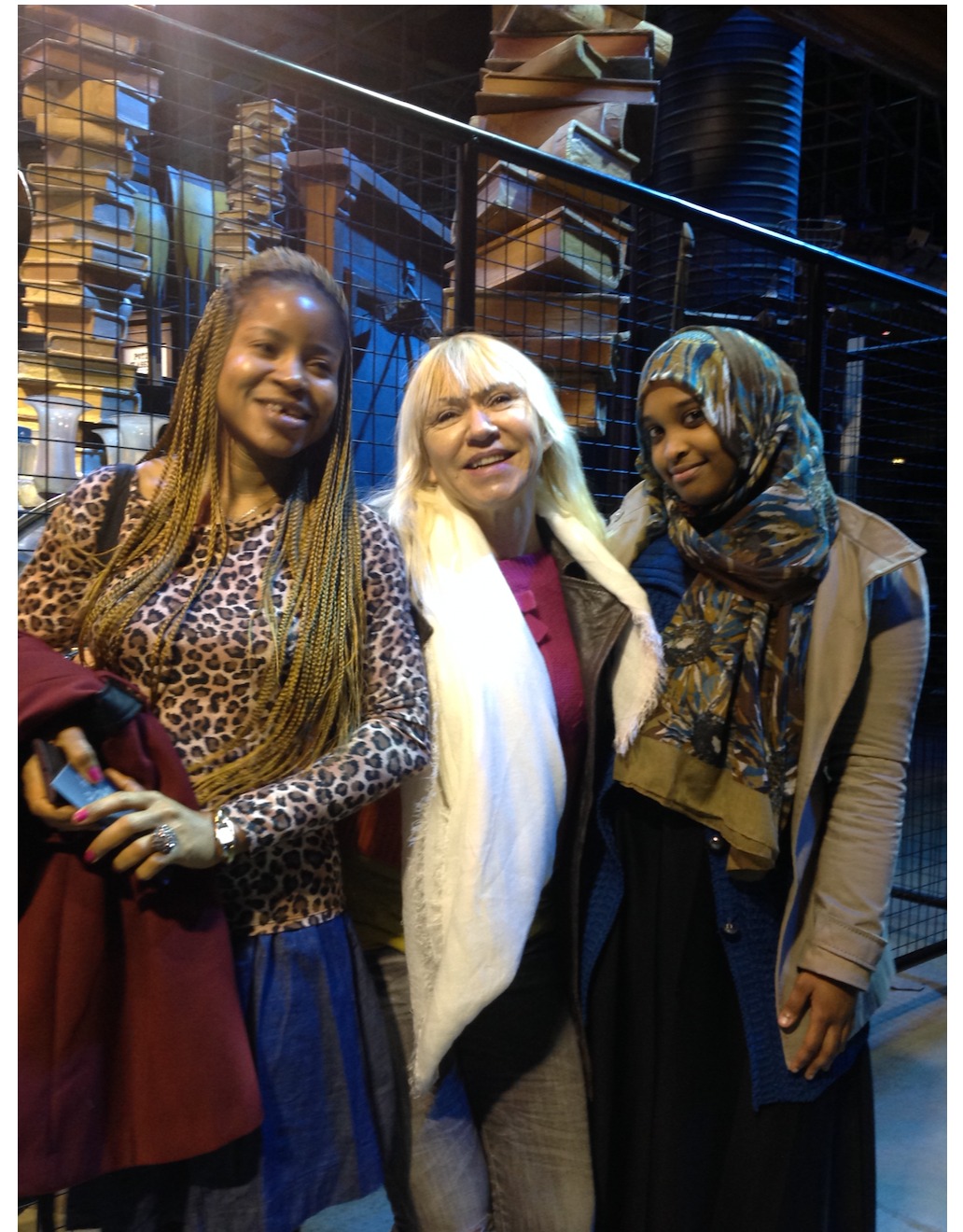 Trzy uśmiechnięte kobiety w różnym wieku, stojące blisko siebie na tle regałów z książkami i metalowej konstrukcji.