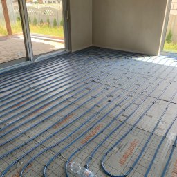 Usługi hydrauliczne - Niebieskie rury ogrzewania podłogowego rozłożone na izolacji z nadrukiem, widok z góry na duży pokój w stanie surowym, butelka wody leży na rurach.