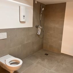 Usługi hydrauliczne - Nowoczesna łazienka z wiszącą toaletą, prysznicem bez brodzika i białym piecem gazowym na ścianie, wykończona szarymi płytkami.