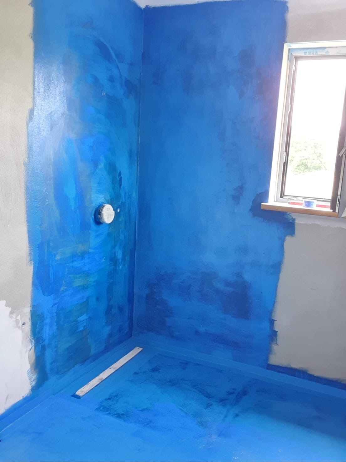 Niebieska hydroizolacja w narożniku łazienki, widoczne ślady po malowaniu na ścianach, okno w tle.