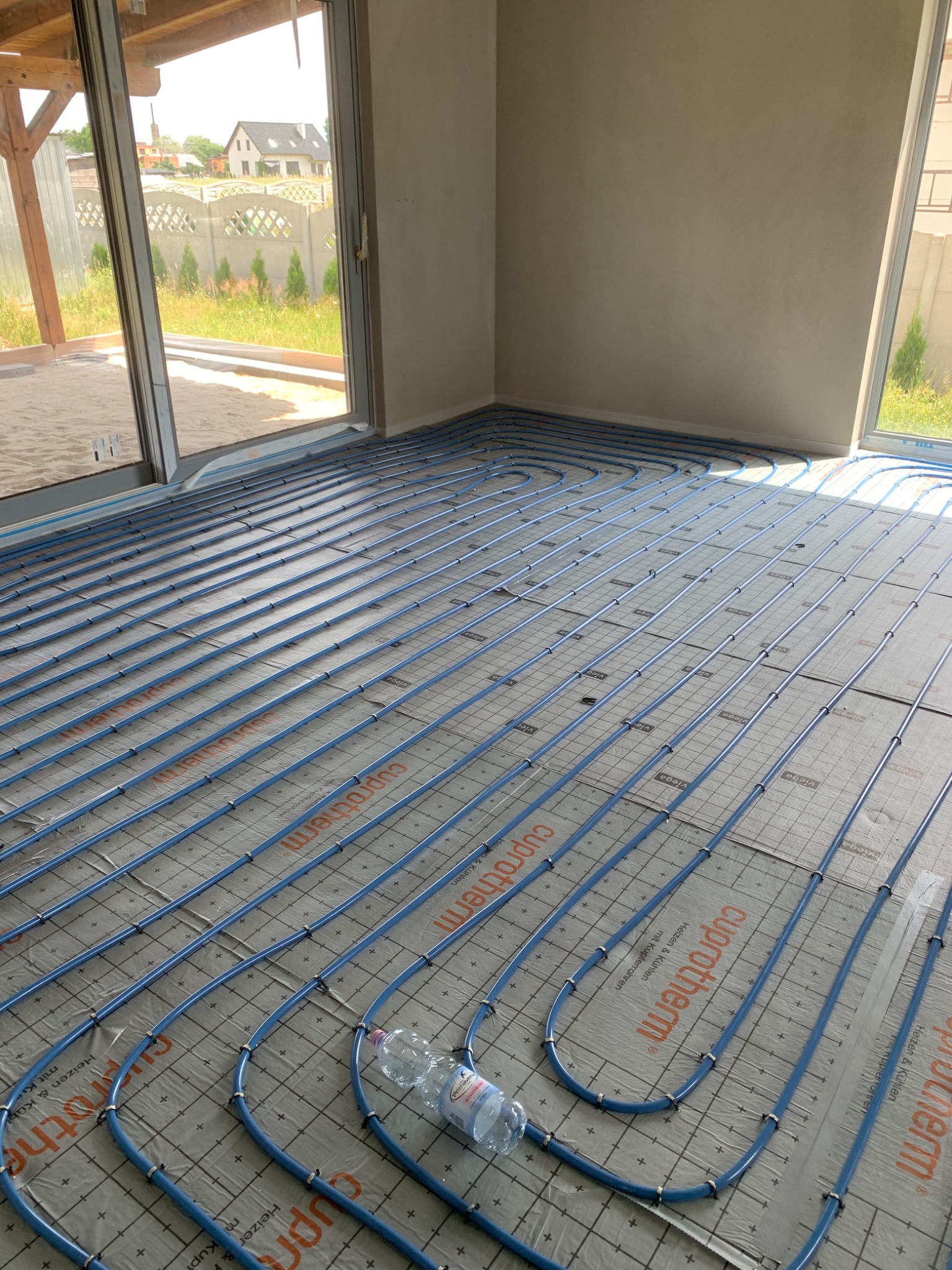 Niebieskie rury ogrzewania podłogowego rozłożone na izolacji z nadrukiem, widok z góry na duży pokój w stanie surowym, butelka wody leży na rurach.