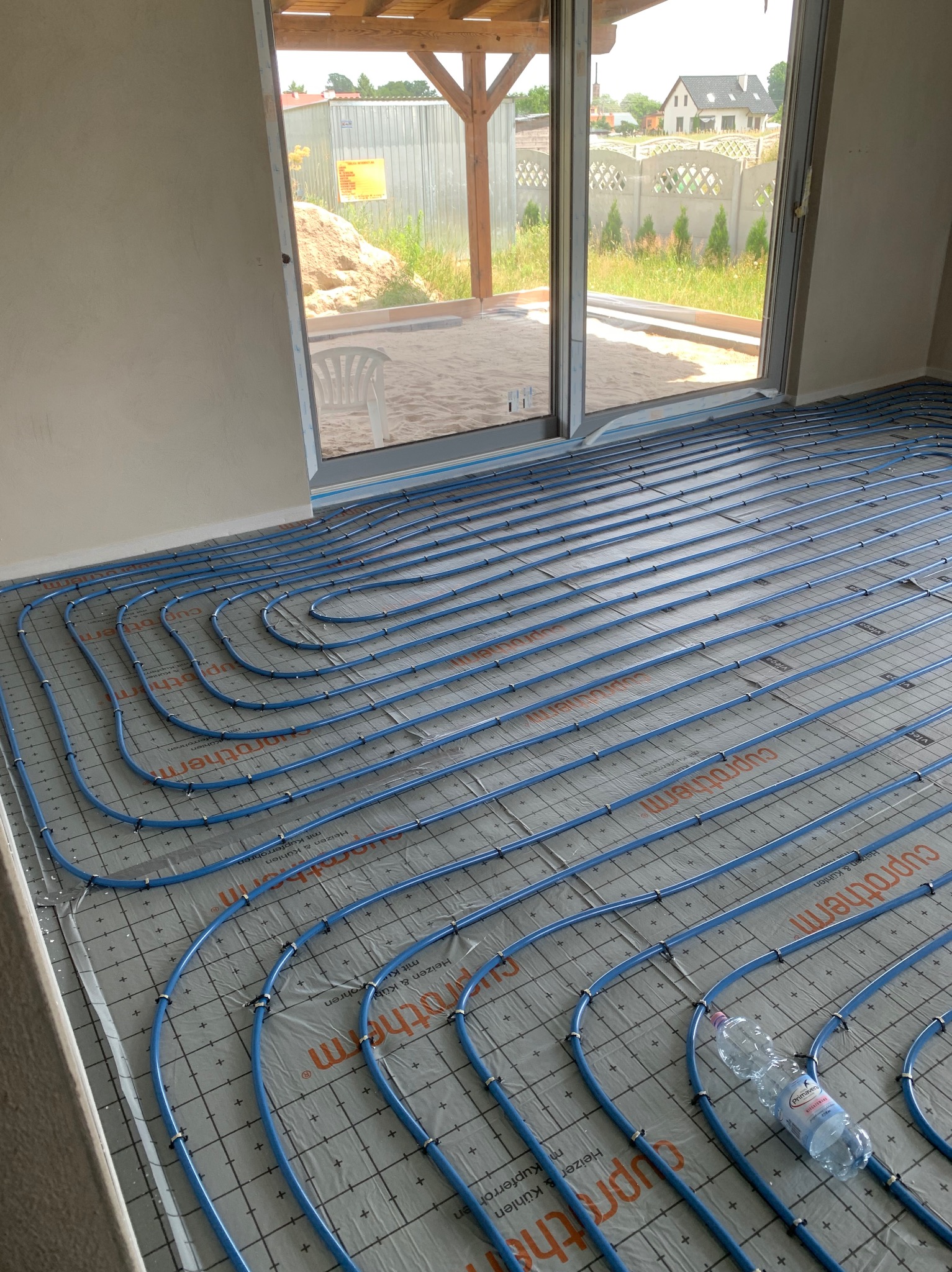 Niebieskie rury ogrzewania podłogowego marki Cuprotherm rozłożone na szarej macie izolacyjnej z widoczną butelką wody mineralnej Primavera, w tle okno z widokiem na ogród i budynek w budowie.