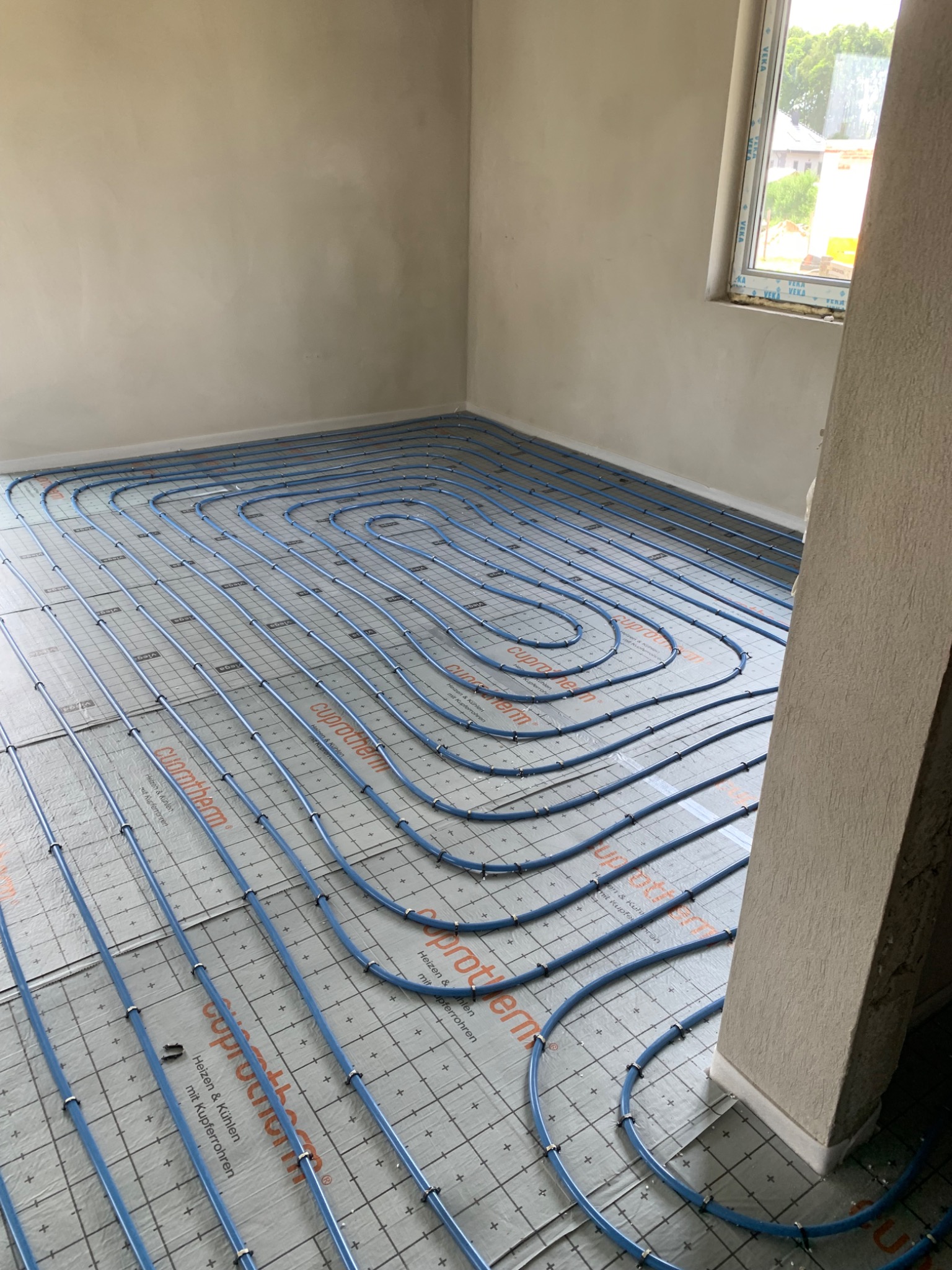 Niebieskie rury ogrzewania podłogowego ułożone spiralnie na izolacji podłogowej w surowym wnętrzu, widoczne mocowania rur i okno w tle.