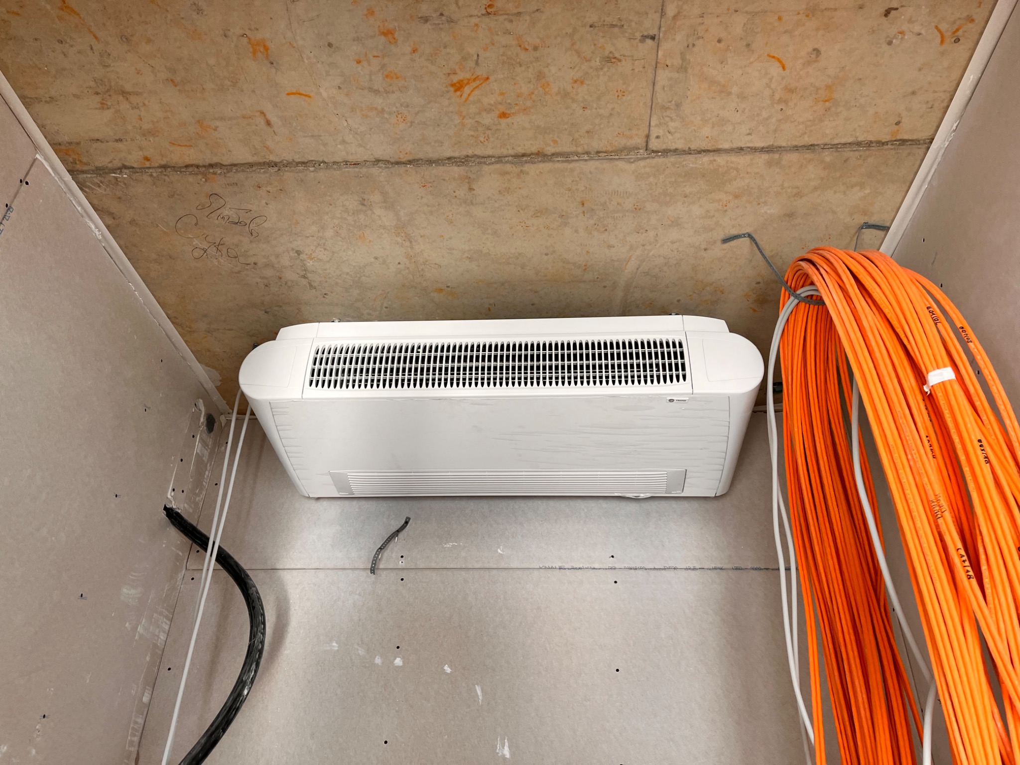 Biały klimakonwektor kasetonowy pod sufitem w surowym wnętrzu, widoczne pomarańczowe kable instalacyjne i czarne rury, ściany w stanie deweloperskim.