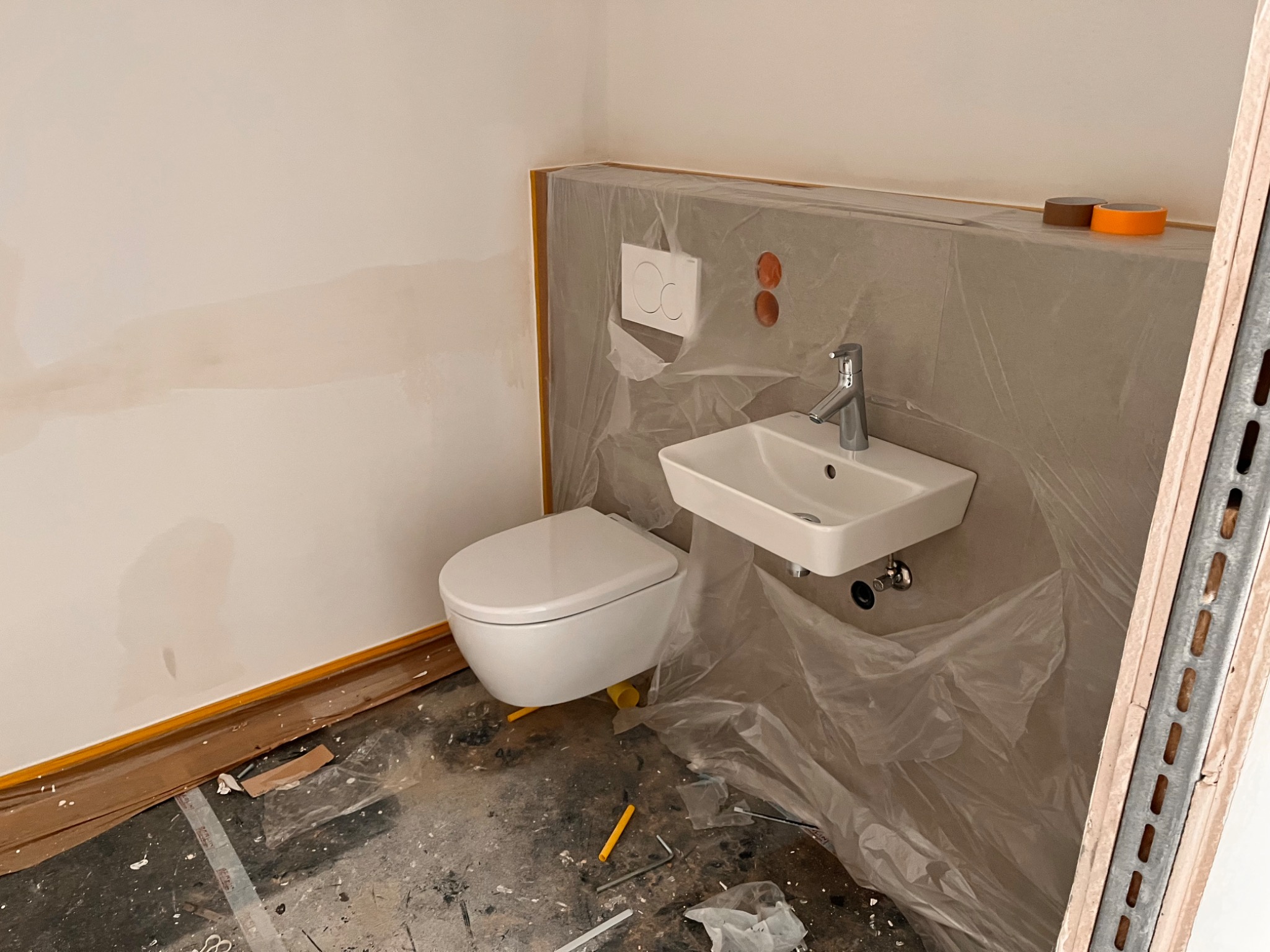 Wisząca toaleta i umywalka zamontowane w łazience w trakcie remontu, zabezpieczone folią ochronną, widoczne narzędzia na podłodze i taśmy malarskie.