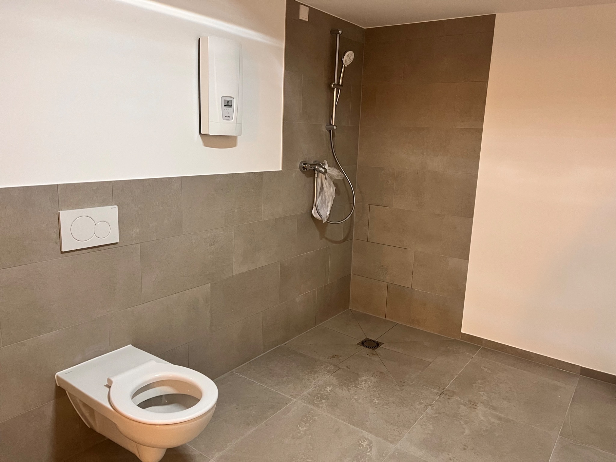 Nowoczesna łazienka z wiszącą toaletą, prysznicem bez brodzika i białym piecem gazowym na ścianie, wykończona szarymi płytkami.