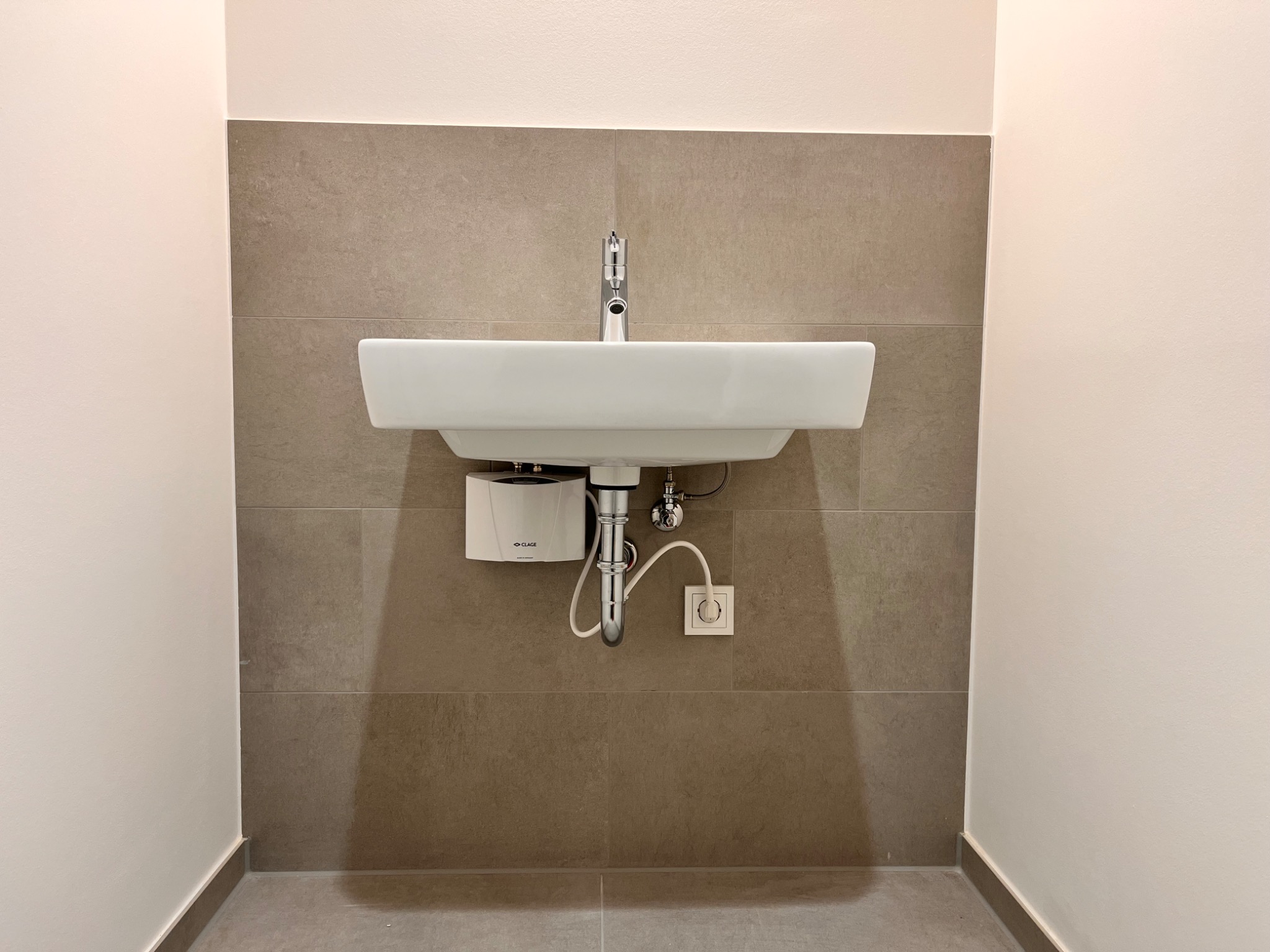 Nowoczesna, prostokątna umywalka z chromowaną baterią zamontowana na ścianie wyłożonej brązowymi płytkami, z widocznym podgrzewaczem wody i gniazdkiem elektrycznym.