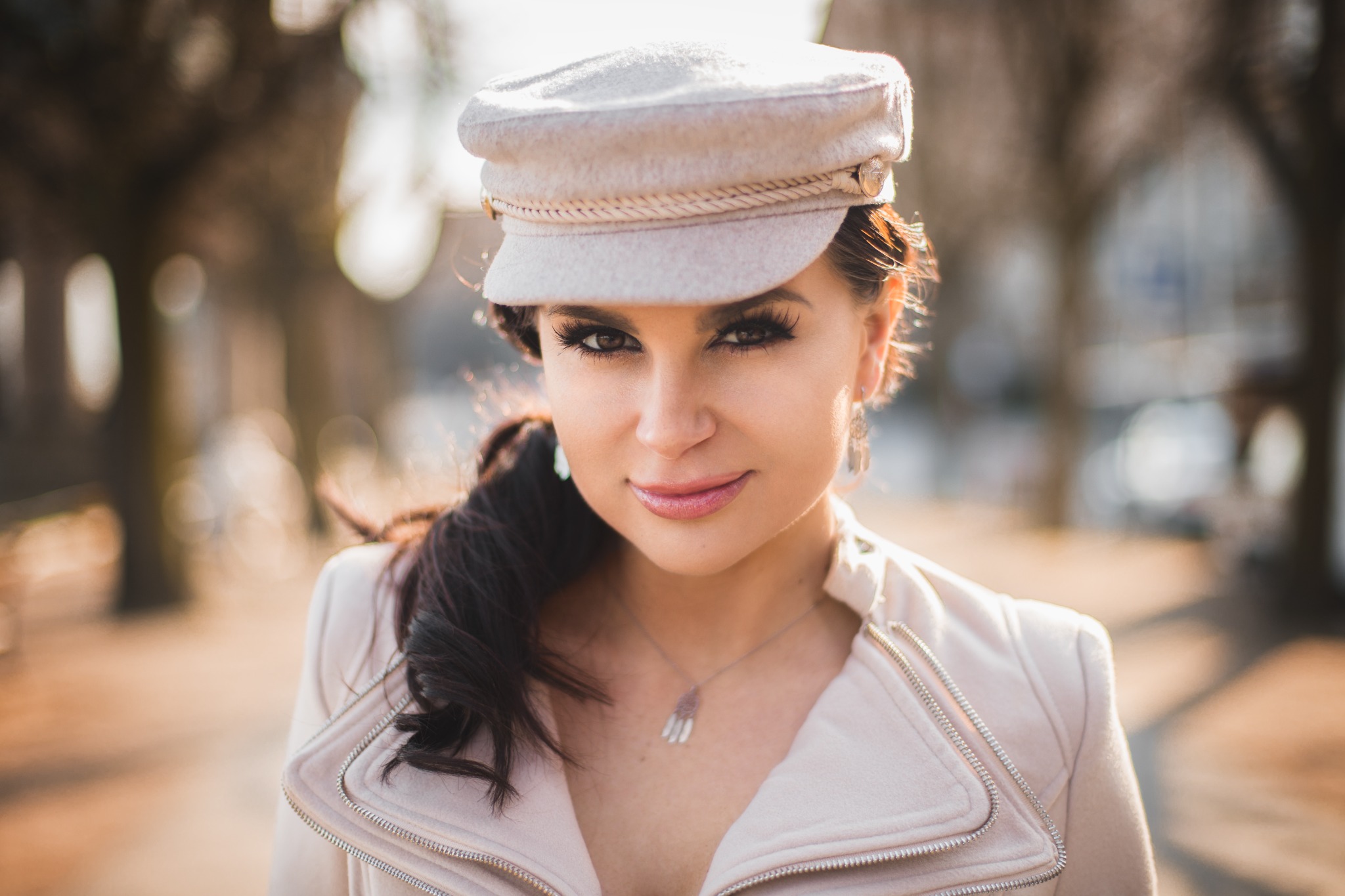 Portret kobiety w jasnym kapeluszu i płaszczu, z delikatnym makijażem i błyszczącą biżuterią, z rozmyciem tła podkreślającym ostrość na twarzy.