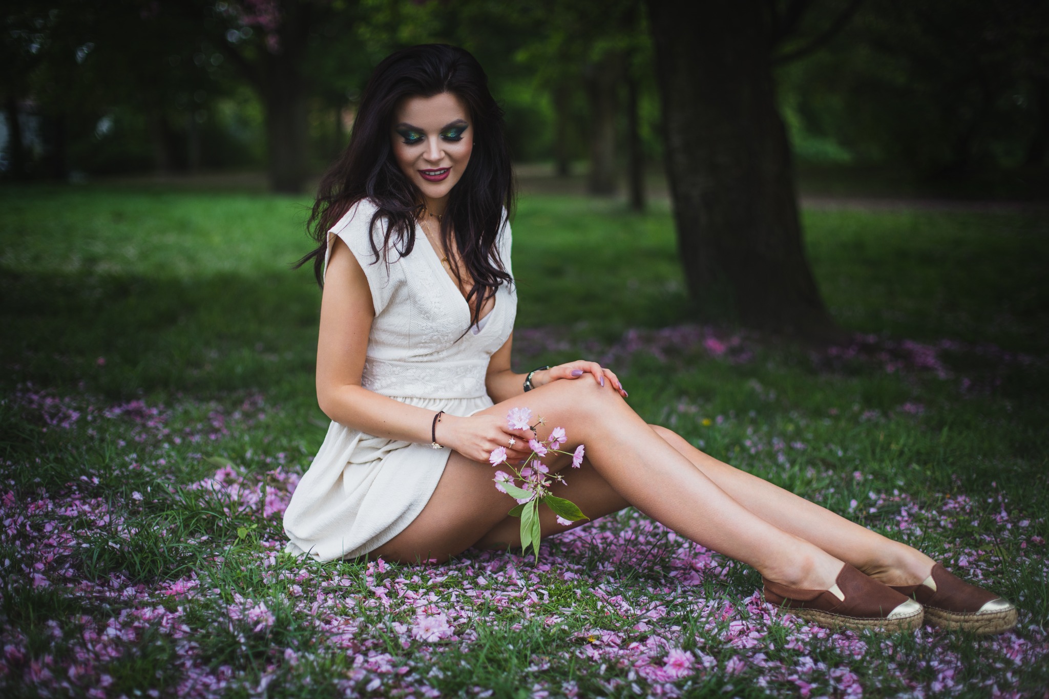 Kobieta w białej sukience siedzi na trawie usłanej różowymi płatkami, trzymając gałązkę kwitnącej wiśni, z ciemnymi włosami i zielonym makijażem oczu.