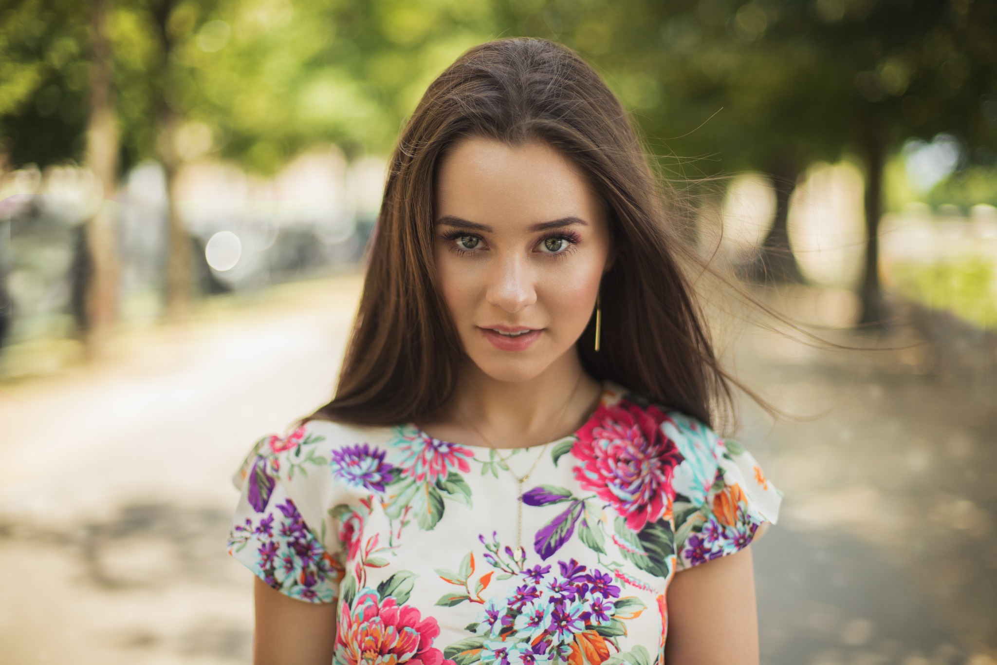 Portret młodej kobiety z długimi brązowymi włosami, w bluzce w kwiaty, z delikatnym makijażem i złotymi kolczykami na tle rozmytego parku.