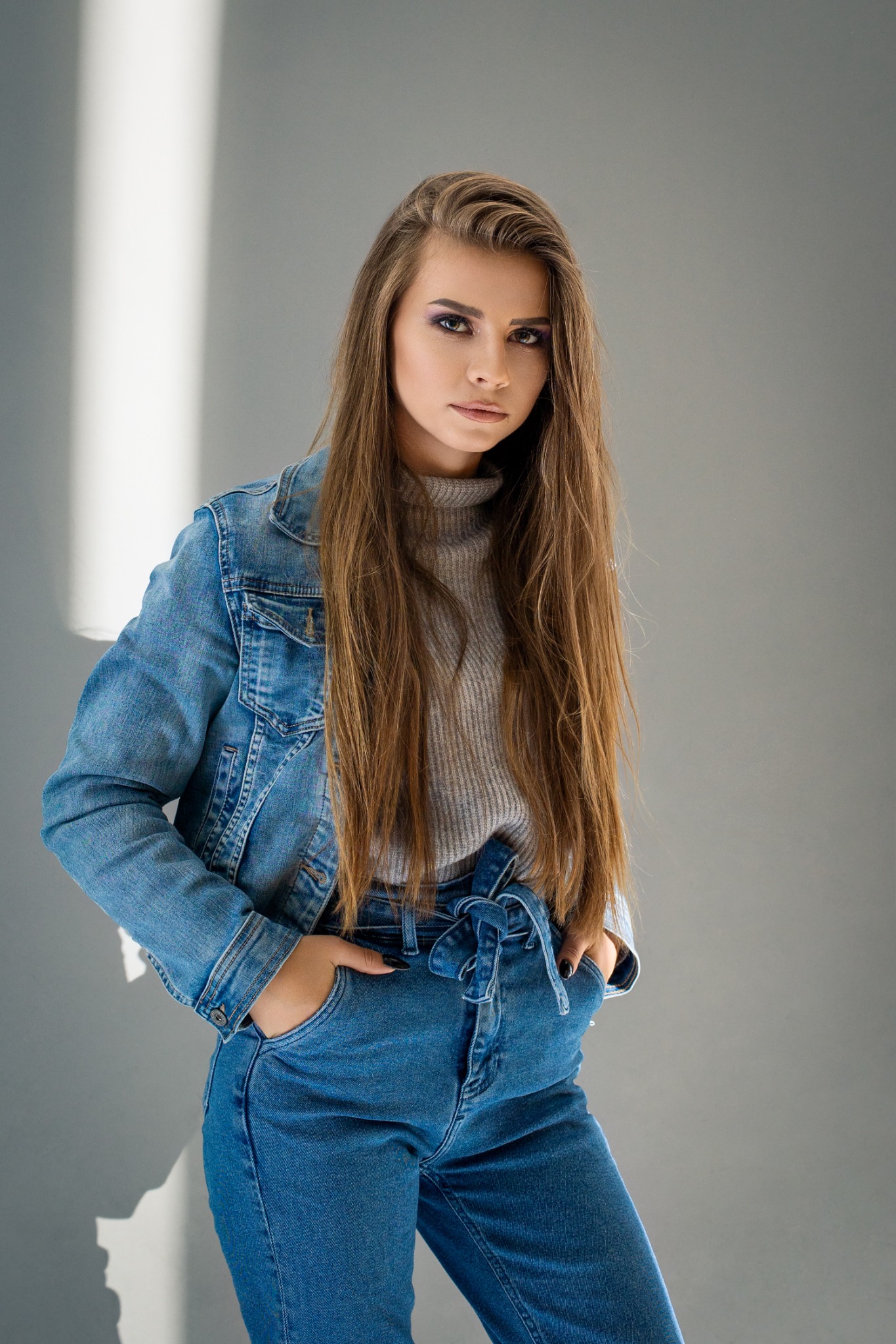 Portret kobiety w dżinsowej kurtce i spodniach z wysokim stanem, pozującej na tle szarej ściany z wyraźnym pasmem światła.