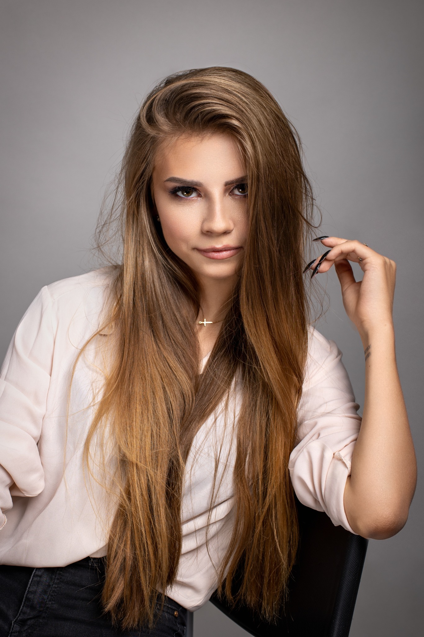 Portret kobiety z długimi brązowymi włosami, w jasnej bluzce i z delikatnym makijażem, siedzącej na krześle na szarym tle.
