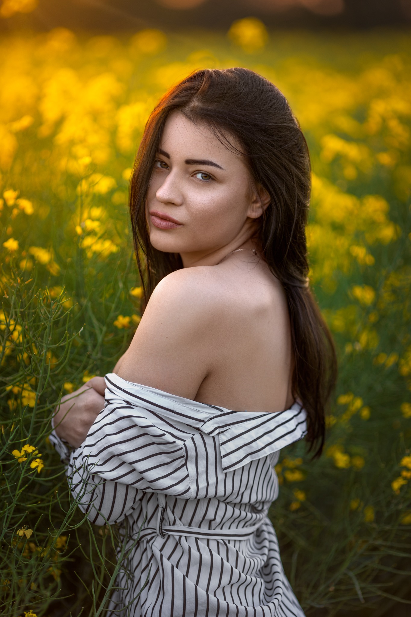 Portret kobiety w koszuli w paski, odwróconej przez ramię, na tle pola kwitnącego rzepaku o zachodzie słońca.