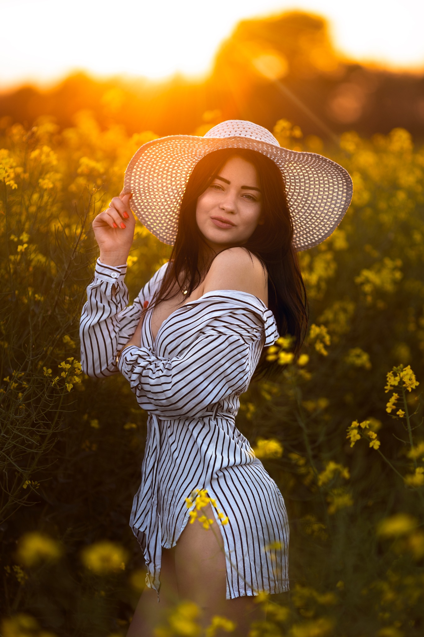 Kobieta w kapeluszu i koszuli w paski pozuje na tle kwitnącego pola rzepaku w ciepłym, zachodzącym słońcu.