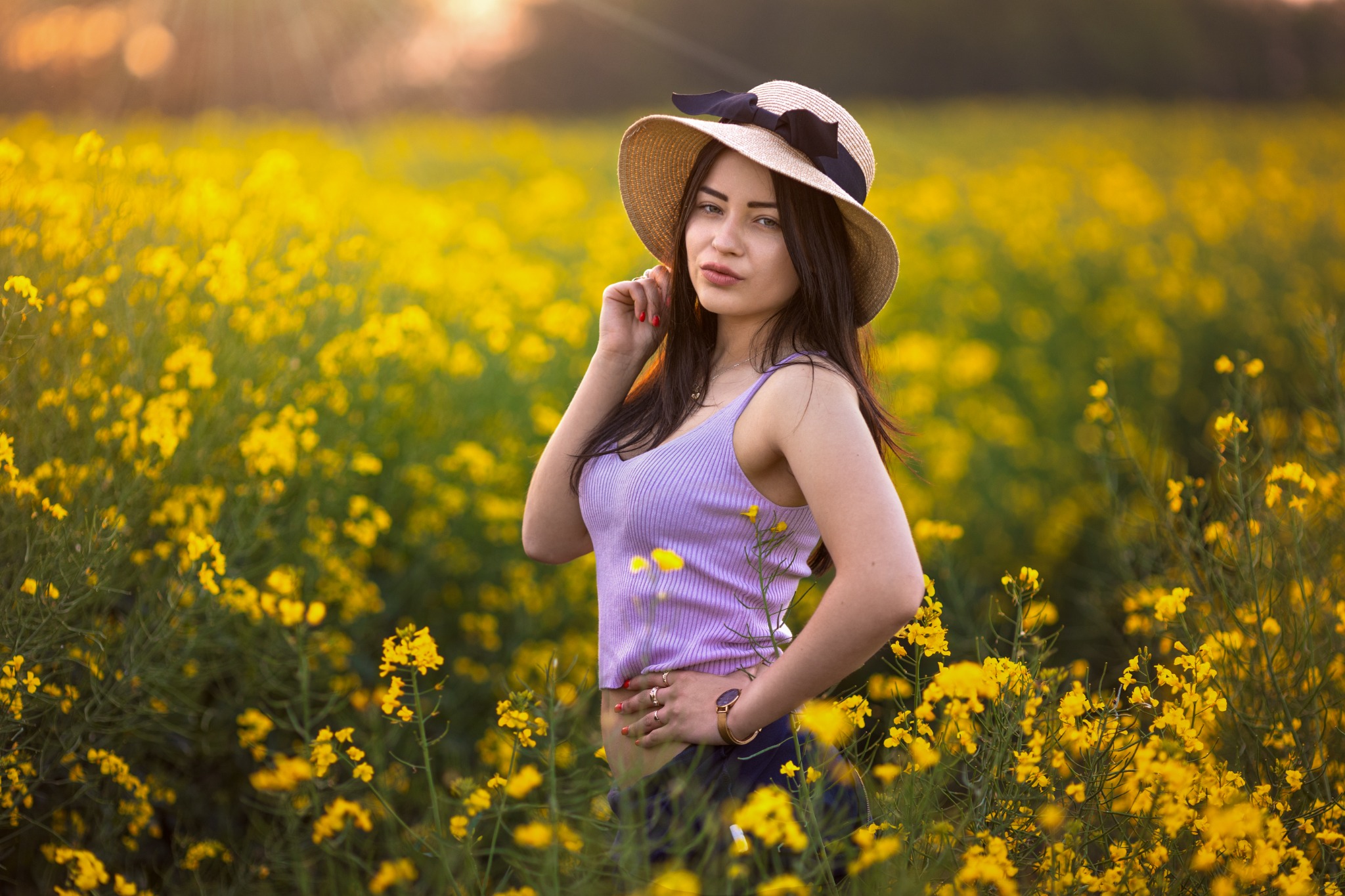 Portret młodej kobiety w słomkowym kapeluszu z czarną wstążką, pozującej na tle pola kwitnącego rzepaku w promieniach zachodzącego słońca.