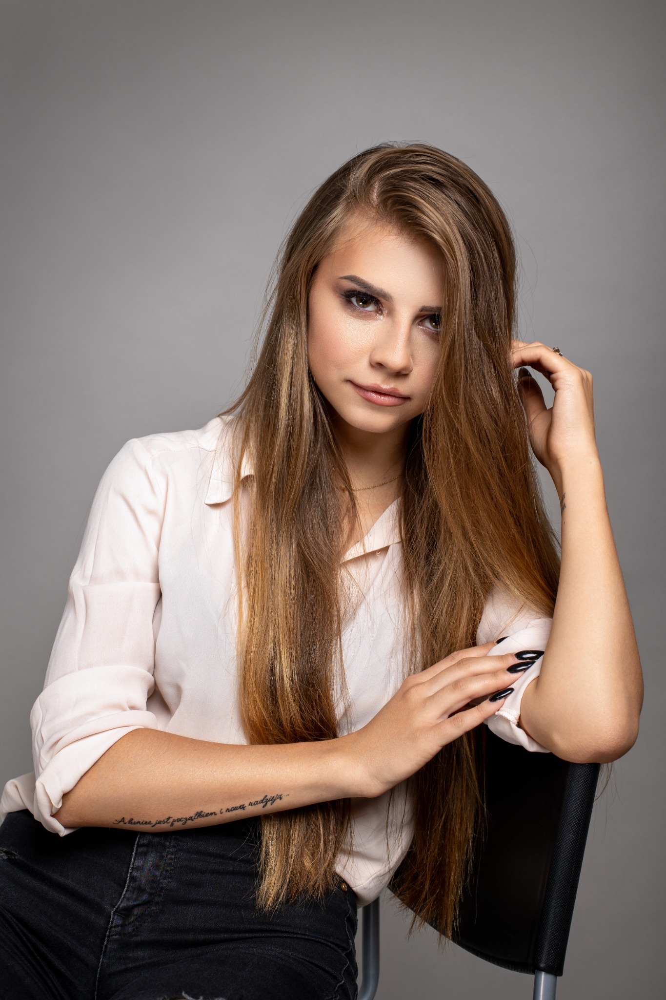 Portret młodej kobiety z długimi, brązowymi włosami, w jasnej koszuli, siedzącej na krześle na szarym tle, z tatuażem na ramieniu i ciemnym makijażem oczu.
