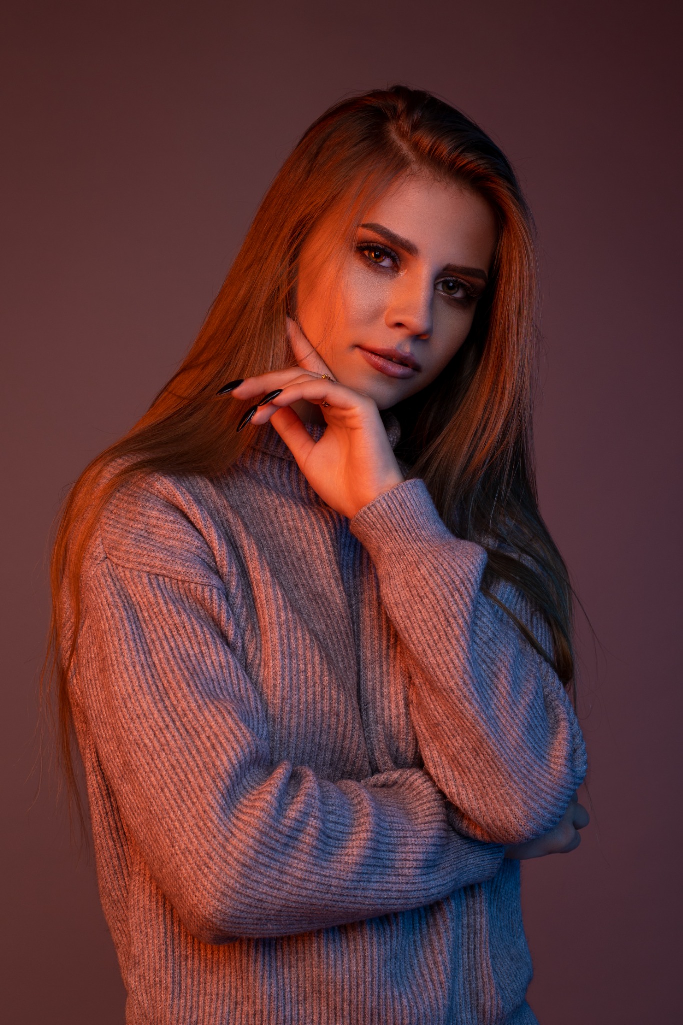 Portret kobiety w szarym swetrze z golfem, oświetlonej pomarańczowo-fioletowym światłem, z dłonią pod brodą i długimi, brązowymi włosami.