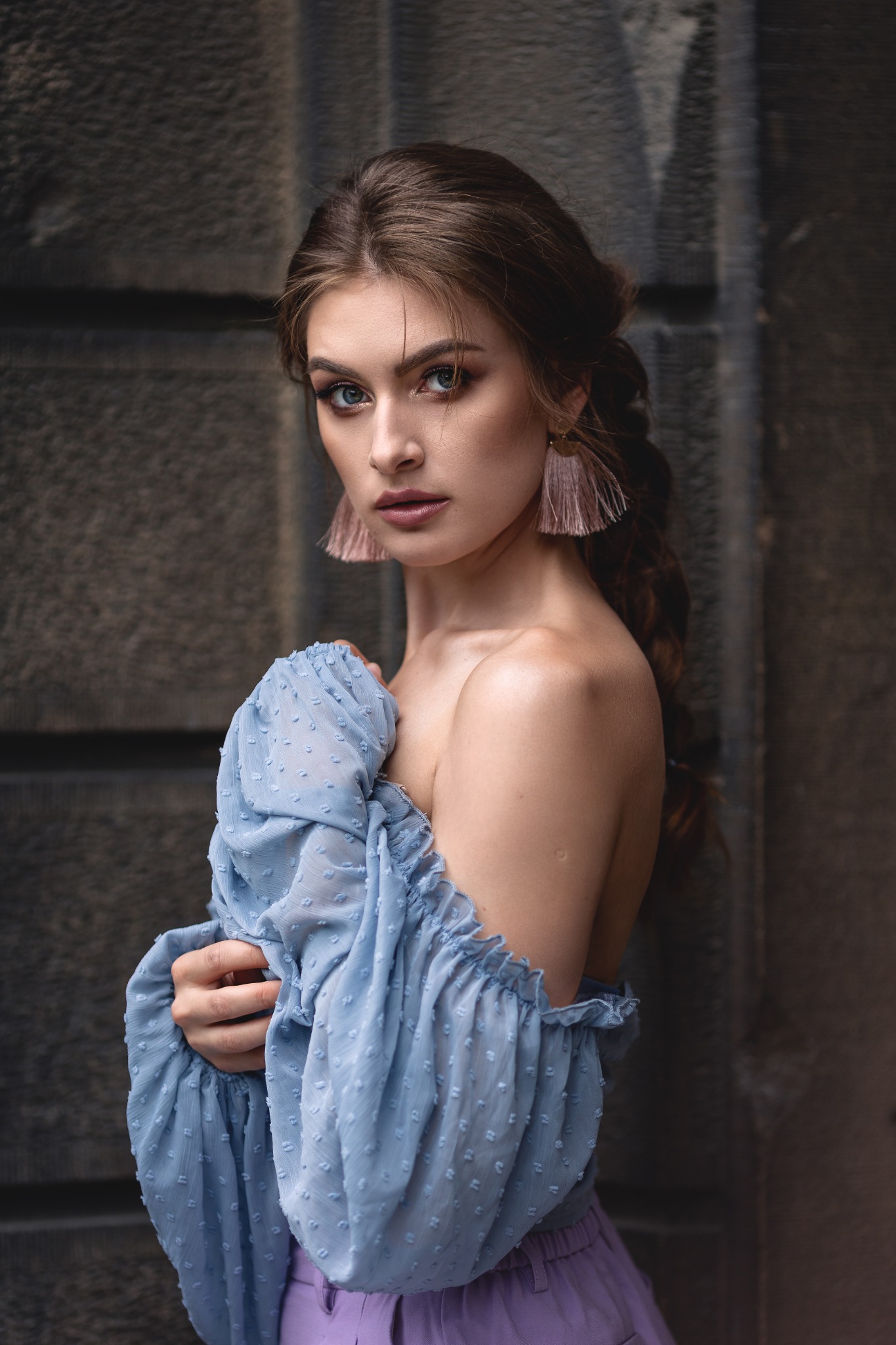 Portret kobiety z warkoczem, w błękitnej bluzce z odkrytymi ramionami i fioletowych spodniach, pozującej na tle szarej, teksturowanej ściany. Uwagę przyciągają długie, pudrowe kolczyki z frędzlami.