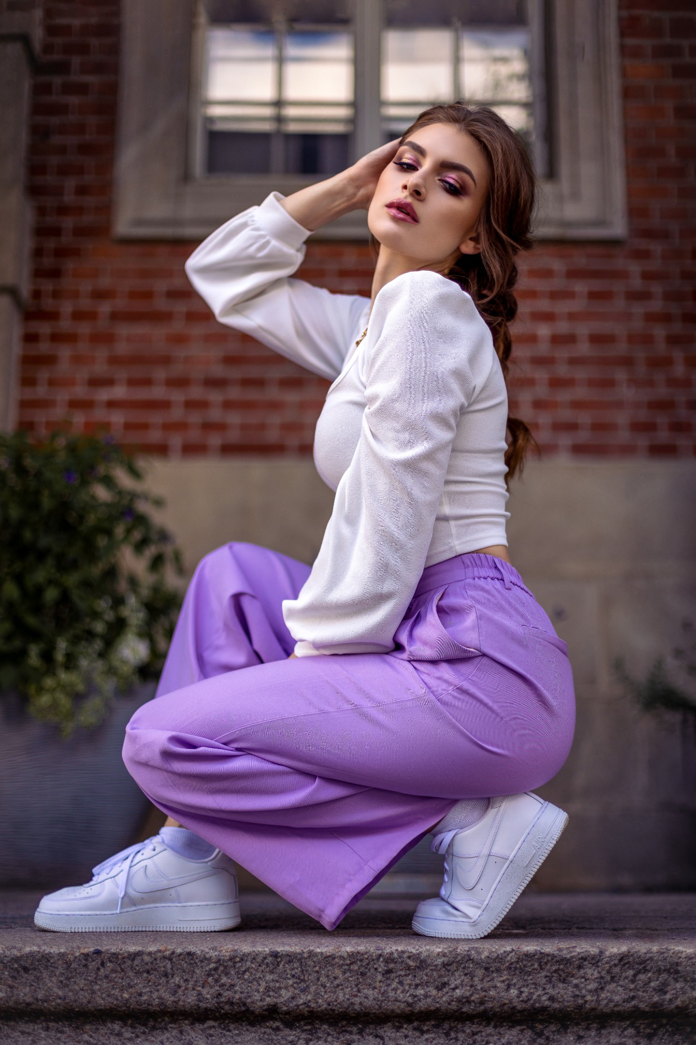 Kobieta w białej bluzce i liliowych spodniach, kuca na schodach, pozując na tle ceglanej ściany z oknem, z ręką uniesioną do włosów i mocnym makijażem.