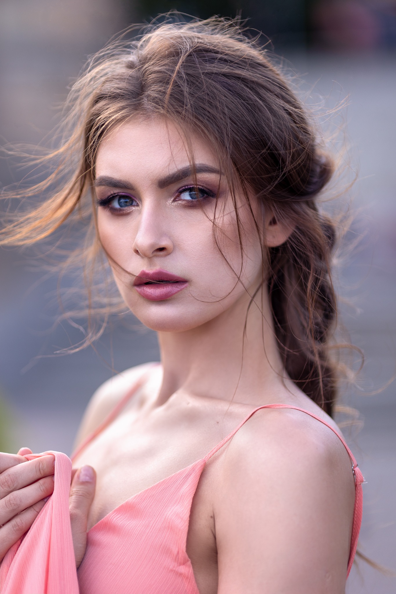 Portret młodej kobiety z rozwianymi włosami, makijażem w odcieniach różu i fioletu, ubrana w delikatną sukienkę na ramiączkach w kolorze brzoskwini, trzymająca materiał sukienki w dłoni.