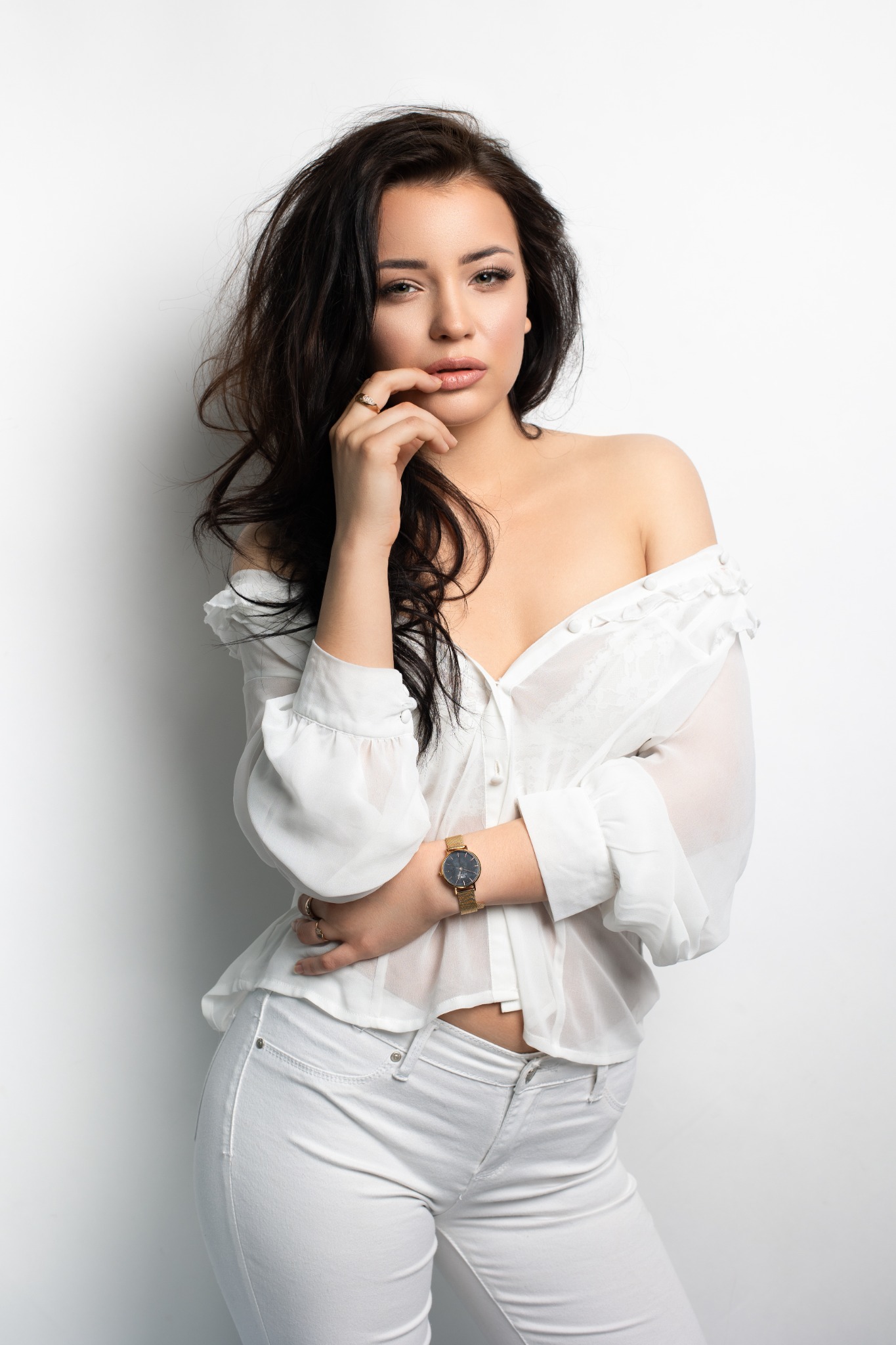Portret kobiety w białej bluzce z odkrytymi ramionami i białych spodniach, z ręką przy twarzy, noszącej złoty zegarek i pierścionki, na jasnym tle.