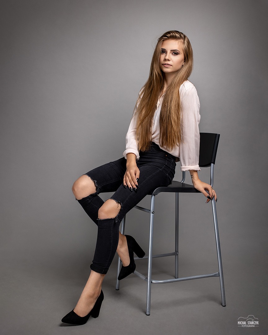 Portret siedzącej kobiety w podartych dżinsach i bluzce z długim rękawem na szarym tle.