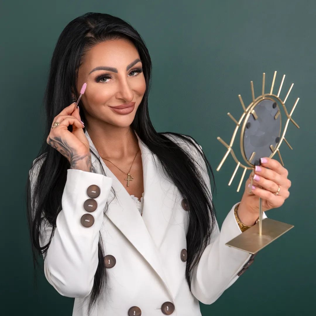 Kobieta w białym żakiecie pozuje z lusterkiem w kształcie oka i szczoteczką do rzęs, prezentując stylizację rzęs na tle ciemnozielonej ściany.