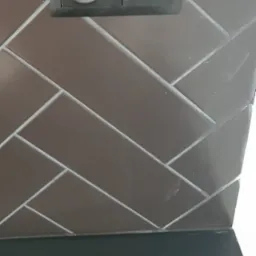 Ułożone w jodełkę ciemnobrązowe płytki ceramiczne z szarą fugą, czarne gniazdko elektryczne i włącznik światła, fragment blatu.