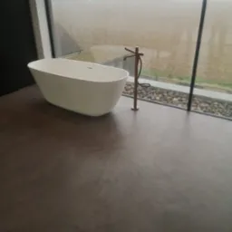 Nowoczesna łazienka z wolnostojącą wanną na tle panoramicznego okna z widokiem na pole w pochmurny dzień. Podłoga wykończona w ciemnoszarym, matowym odcieniu.