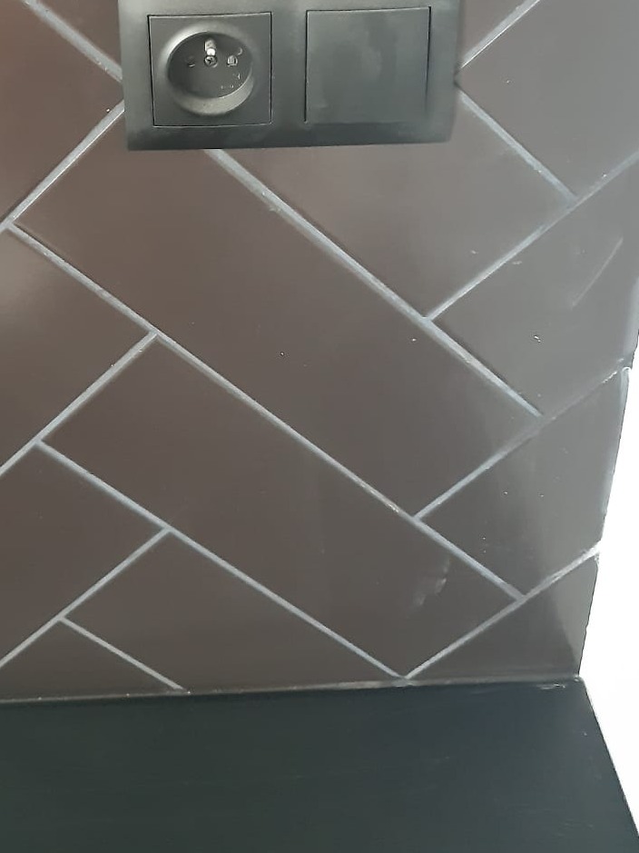 Ułożone w jodełkę ciemnobrązowe płytki ceramiczne z szarą fugą, czarne gniazdko elektryczne i włącznik światła, fragment blatu.