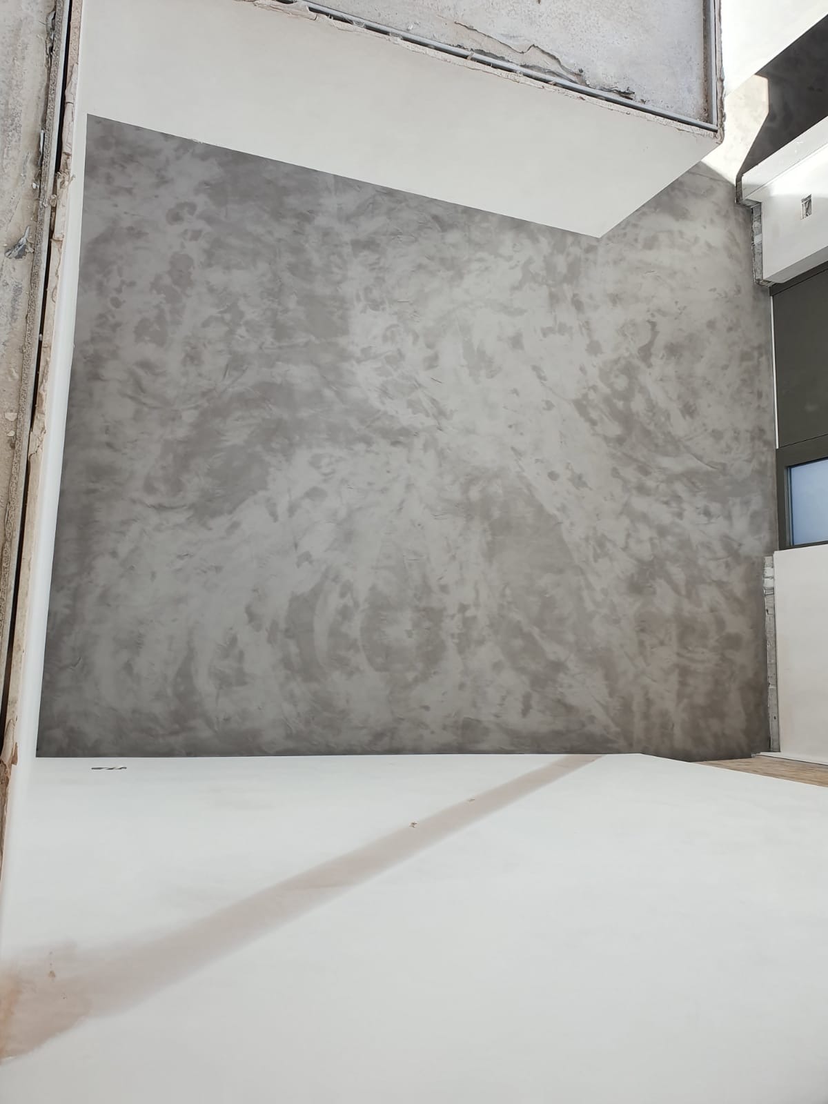 Ściana w surowym, szarym wykończeniu imitującym beton, z widoczną strukturą i delikatnymi smugami, kontrastująca z białą podłogą w surowym stanie.