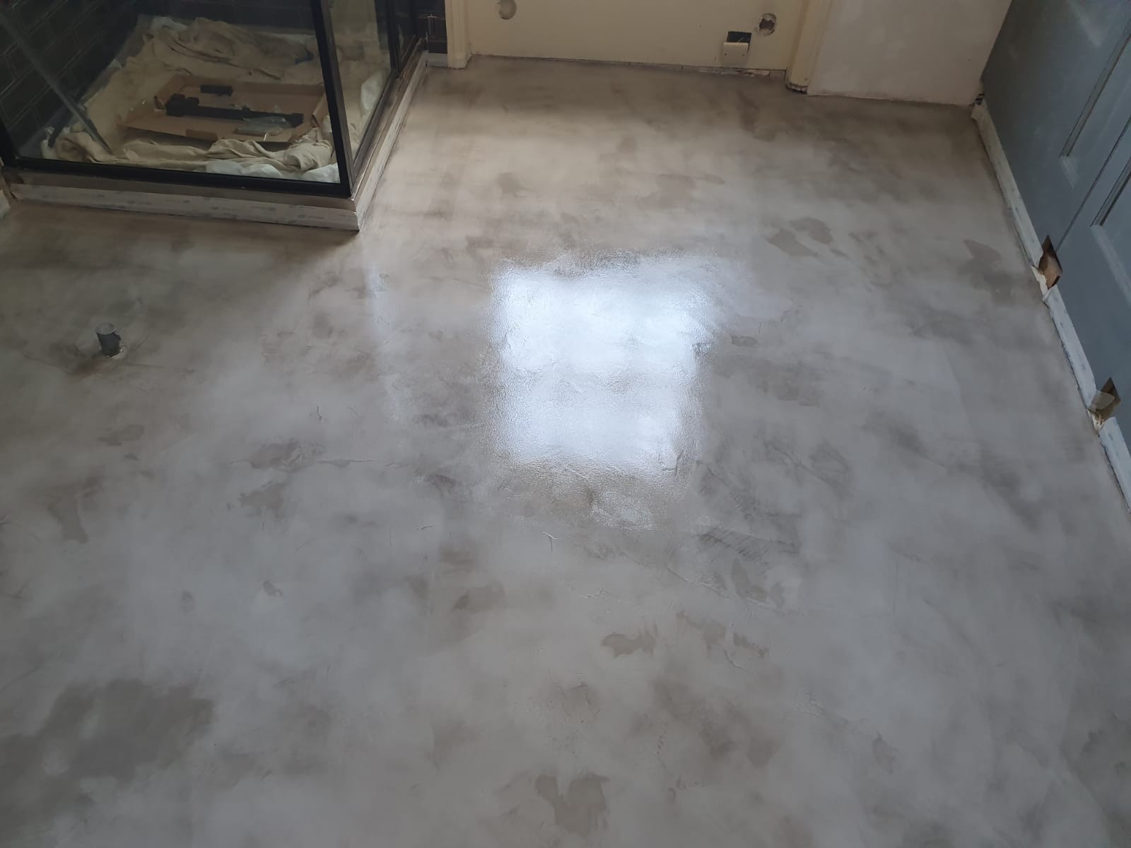 Wypoziomowana, jasnoszara posadzka z efektem chmurkowania, świeżo po wylaniu, z widocznym odbiciem światła, niedokończone wykończenie ściany i drzwi w tle.