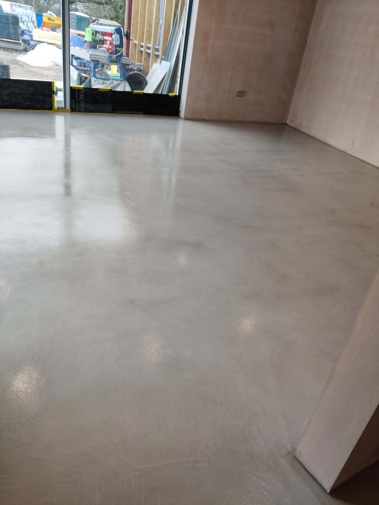 Wypoziomowana, jasnoszara wylewka samopoziomująca w nowym pomieszczeniu z surowymi ścianami i dużym oknem z widokiem na plac budowy.