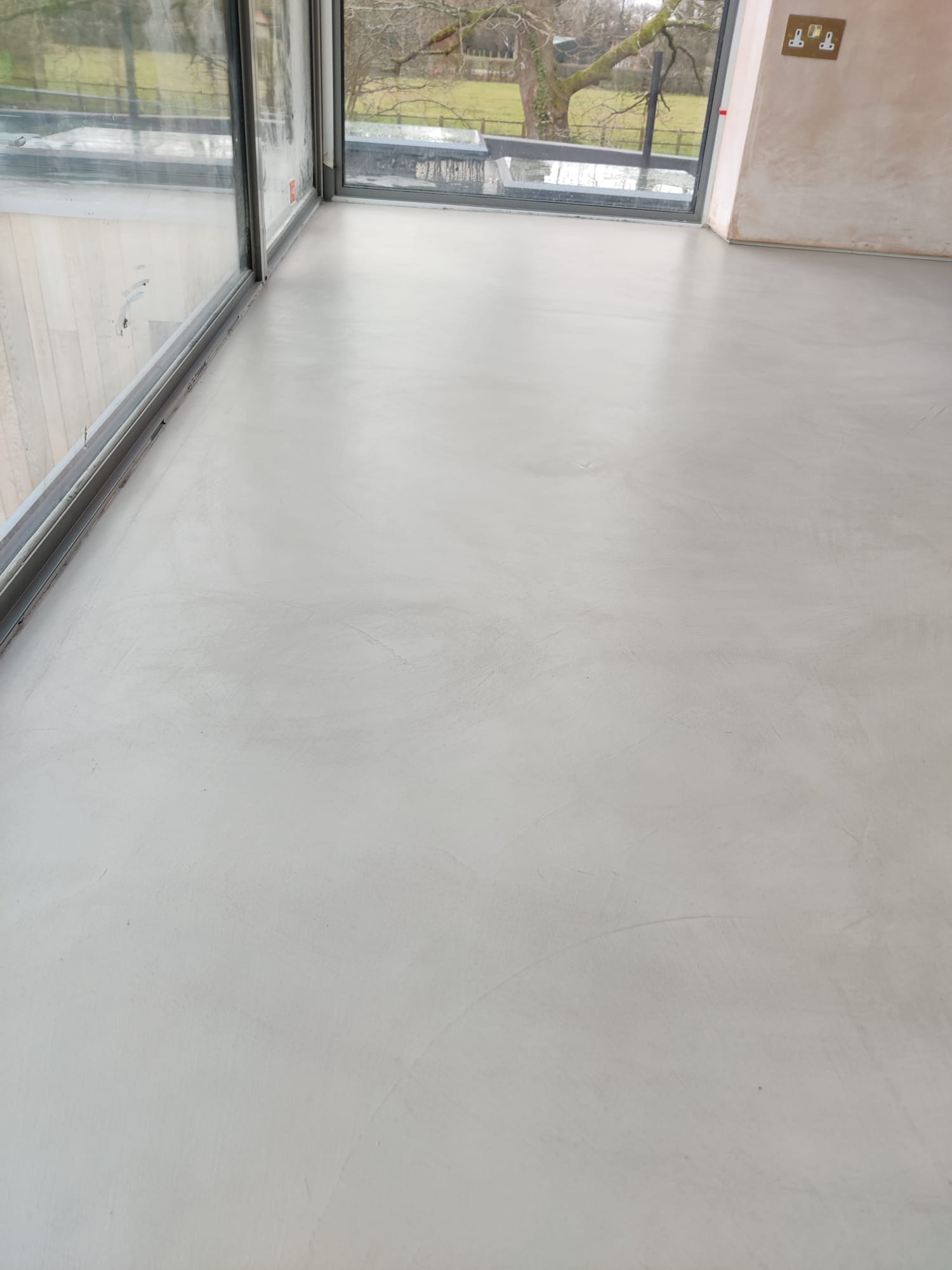 Wnętrze pokoju z wygładzoną, jasnoszarą posadzką żywiczną, duże okno z widokiem na zieleń, ściany w trakcie prac wykończeniowych z widocznymi gniazdkami elektrycznymi.