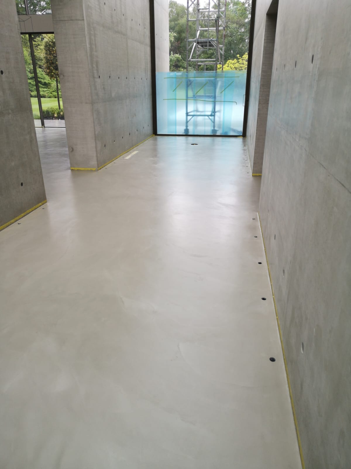 Wnętrze modernistycznego budynku z szarą, gładką posadzką. Żółta taśma zabezpieczająca na styku podłogi i ścian. Widoczne betonowe ściany i filary. W tle okno z niebieską folią i rusztowaniem...