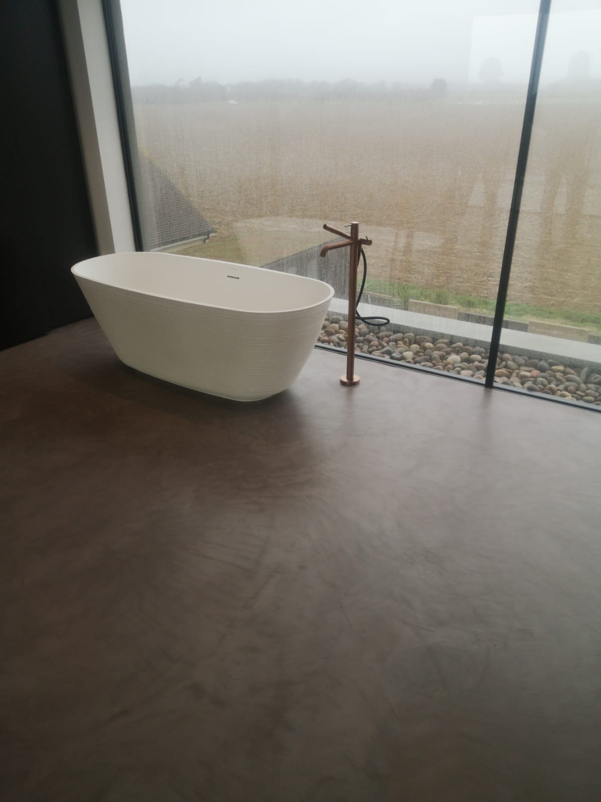 Nowoczesna łazienka z wolnostojącą wanną na tle panoramicznego okna z widokiem na pole w pochmurny dzień. Podłoga wykończona w ciemnoszarym, matowym odcieniu.