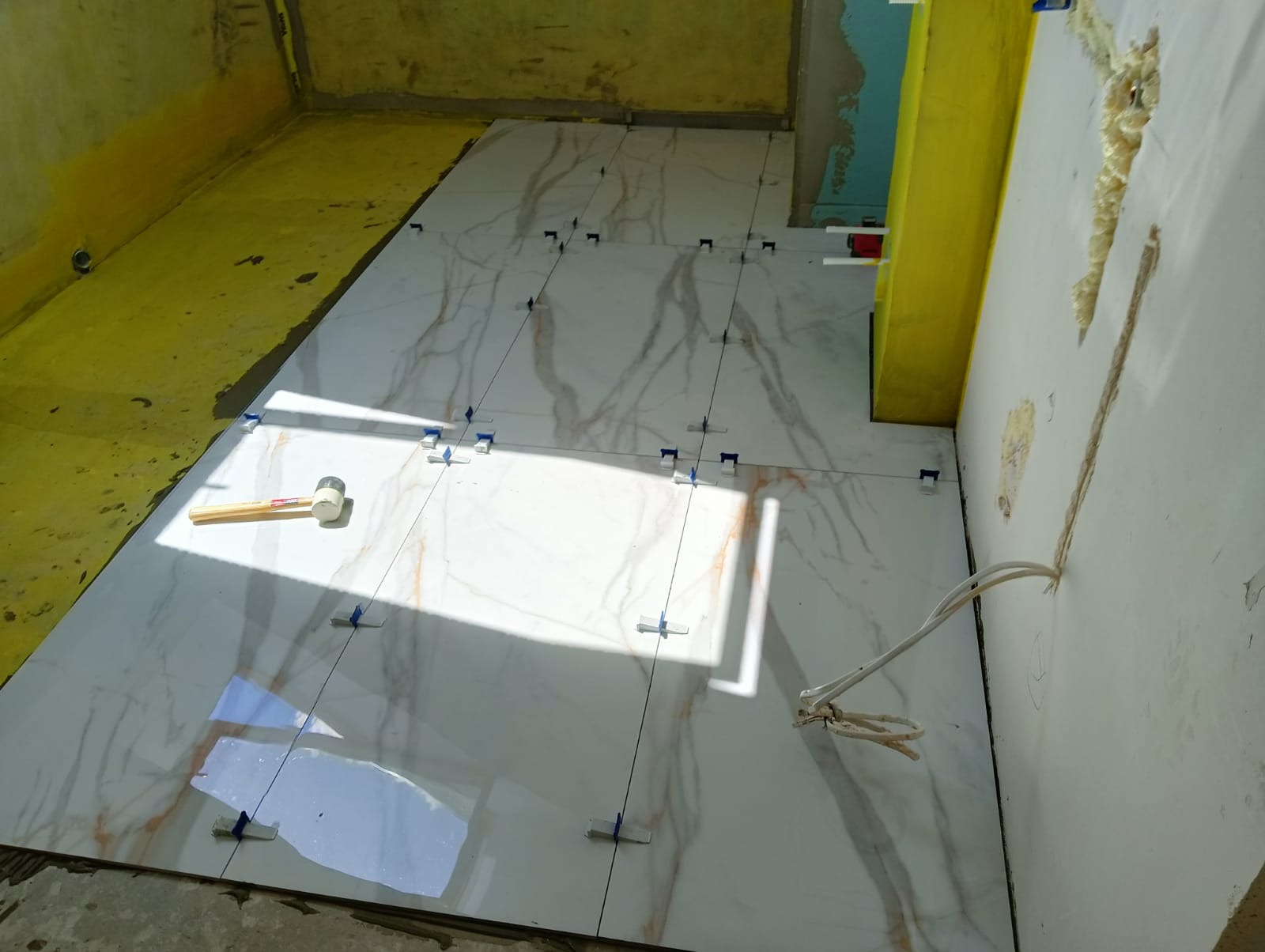 Układanie białych płytek podłogowych z marmurowym wzorem, używając niebieskich klipsów poziomujących i gumowego młotka. Widoczne przewody elektryczne wystające ze ściany i żółta hydroizolacja...