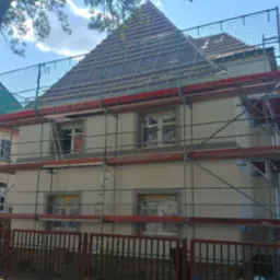 Dwupiętrowy dom w trakcie prac elewacyjnych, z rusztowaniem i siatką ochronną, okna zabezpieczone folią, widoczne fragmenty dachu i drzew