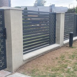 ART PROCOLOR SPÓŁKA Z OGRANICZONĄ ODPOWIEDZIALNOŚCIĄ - Nowoczesne ogrodzenie z metalowymi przęsłami w kolorze grafitowym, z elementami dekoracyjnymi w postaci ażurowych paneli z motywem geometrycznym, osadzone na betonowych słupkach i podmurówce, widok...