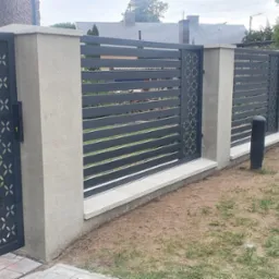 Nowoczesne ogrodzenie z metalowymi przęsłami w kolorze grafitowym, z elementami dekoracyjnymi w postaci ażurowych paneli z motywem geometrycznym, osadzone na betonowych słupkach i podmur&oacute;wce, widok...