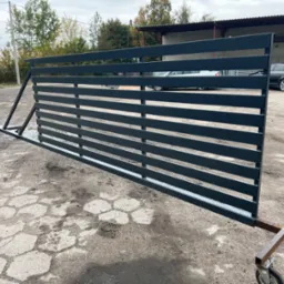 Metalowa brama przesuwna w kolorze grafitowym, z poziomymi profilami, zamontowana na tymczasowych wózkach z kółkami, prezentowana na zewnątrz w szarym otoczeniu przemysłowym.