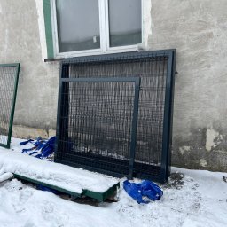 ART PROCOLOR SPÓŁKA Z OGRANICZONĄ ODPOWIEDZIALNOŚCIĄ - Dwa panele ogrodzeniowe w kolorze zielonym i ciemnoszarym, o kratowej strukturze, oparte o szarą ścianę budynku w zimowej scenerii. Na ziemi leżą fragmenty konstrukcji przykryte śniegiem...