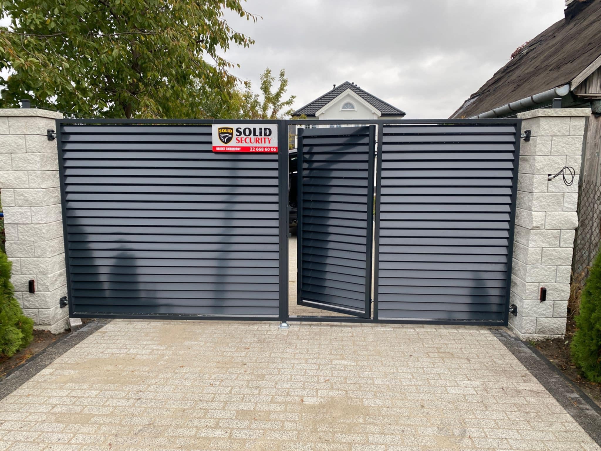 Nowoczesna, grafitowa brama wjazdowa z poziomymi lamelami, częściowo otwarta, zamontowana na murowanych słupkach z jasnego kamienia, z widocznym logo firmy ochroniarskiej i numerem telefonu...