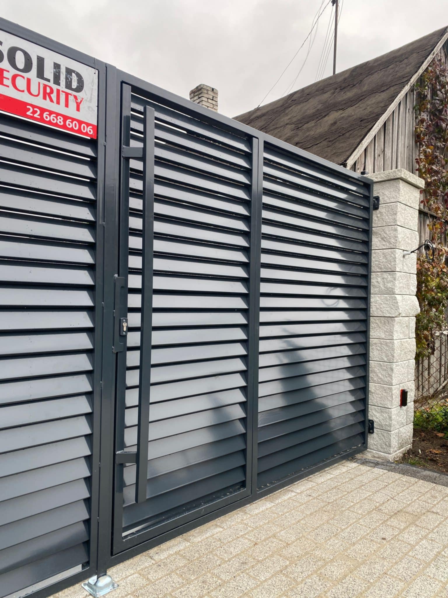 Nowoczesna, szara brama wjazdowa z poziomymi lamelami, zamontowana na słupku z jasnego kamienia, z widocznym szyldem firmy ochroniarskiej.