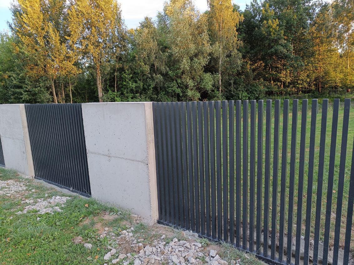 Nowoczesne ogrodzenie z betonowymi słupkami i metalowymi przęsłami pionowymi, widoczne na tle drzew.