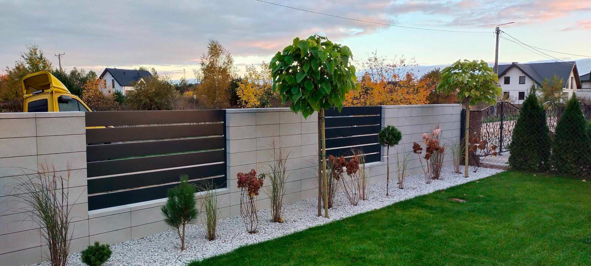 Nowoczesne ogrodzenie z betonowych bloczków i poziomych, ciemnych paneli, uzupełnione ozdobnymi drzewkami i krzewami, z widocznym fragmentem bramy w tle i żółtym samochodem dostawczym za ogrodzeniem.