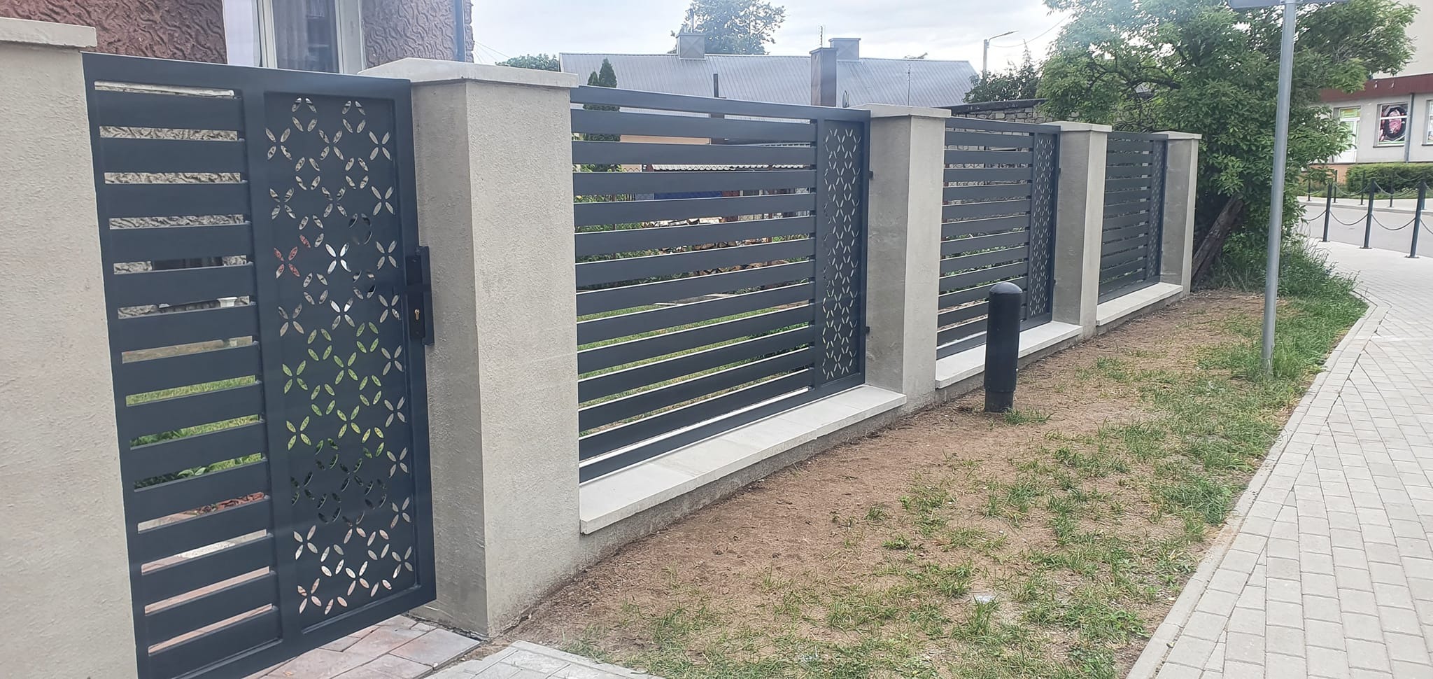 Nowoczesne ogrodzenie z metalowymi przęsłami w kolorze grafitowym, z elementami dekoracyjnymi w postaci ażurowych paneli z motywem geometrycznym, osadzone na betonowych słupkach i podmurówce, widok...