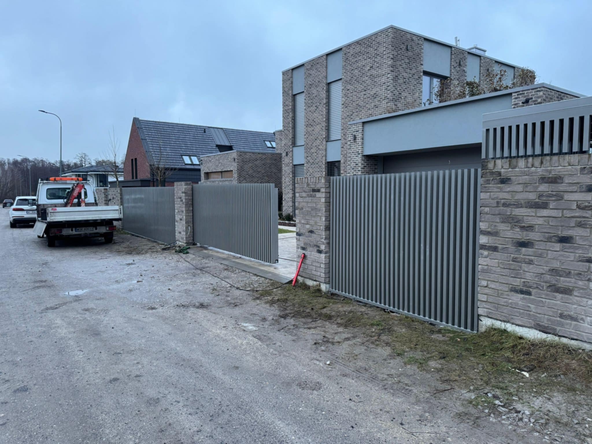 Nowoczesna posesja z przesuwną bramą wjazdową z pionowych profili, dopasowaną kolorystycznie do elewacji domu z szarej cegły, widoczny samochód dostawczy z tyłu.