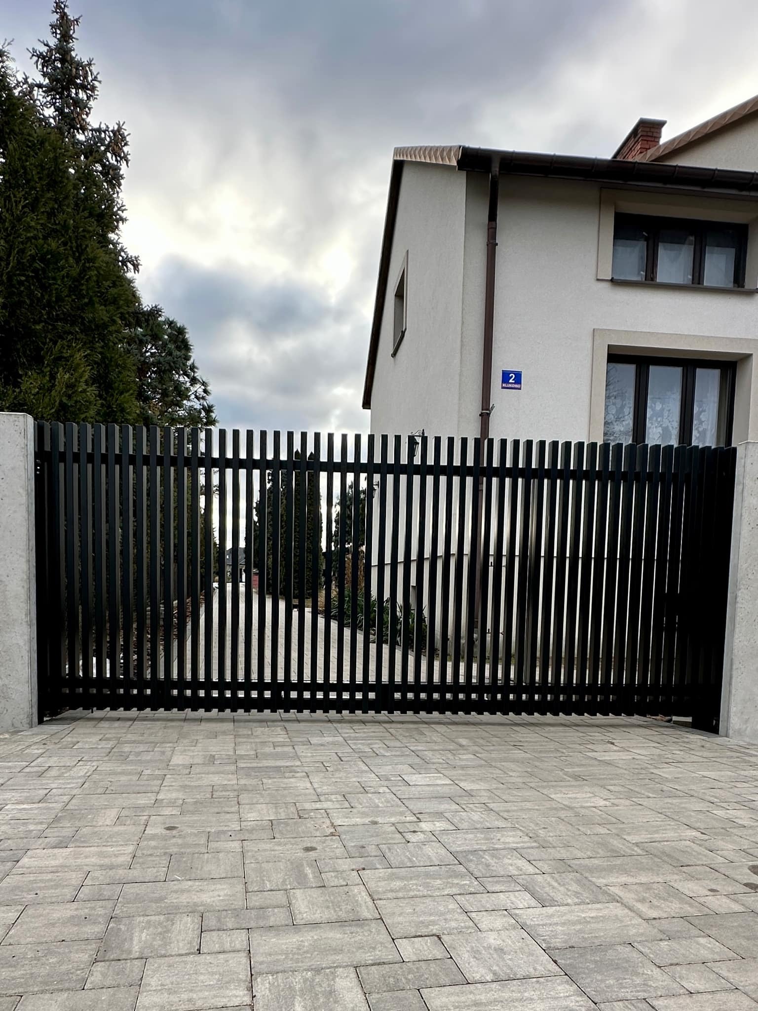 Nowoczesna, czarna brama wjazdowa z pionowych profili, osadzona na betonowych słupach, widok z zewnątrz na tle domu z numerem 2 i brukowanego podjazdu.
