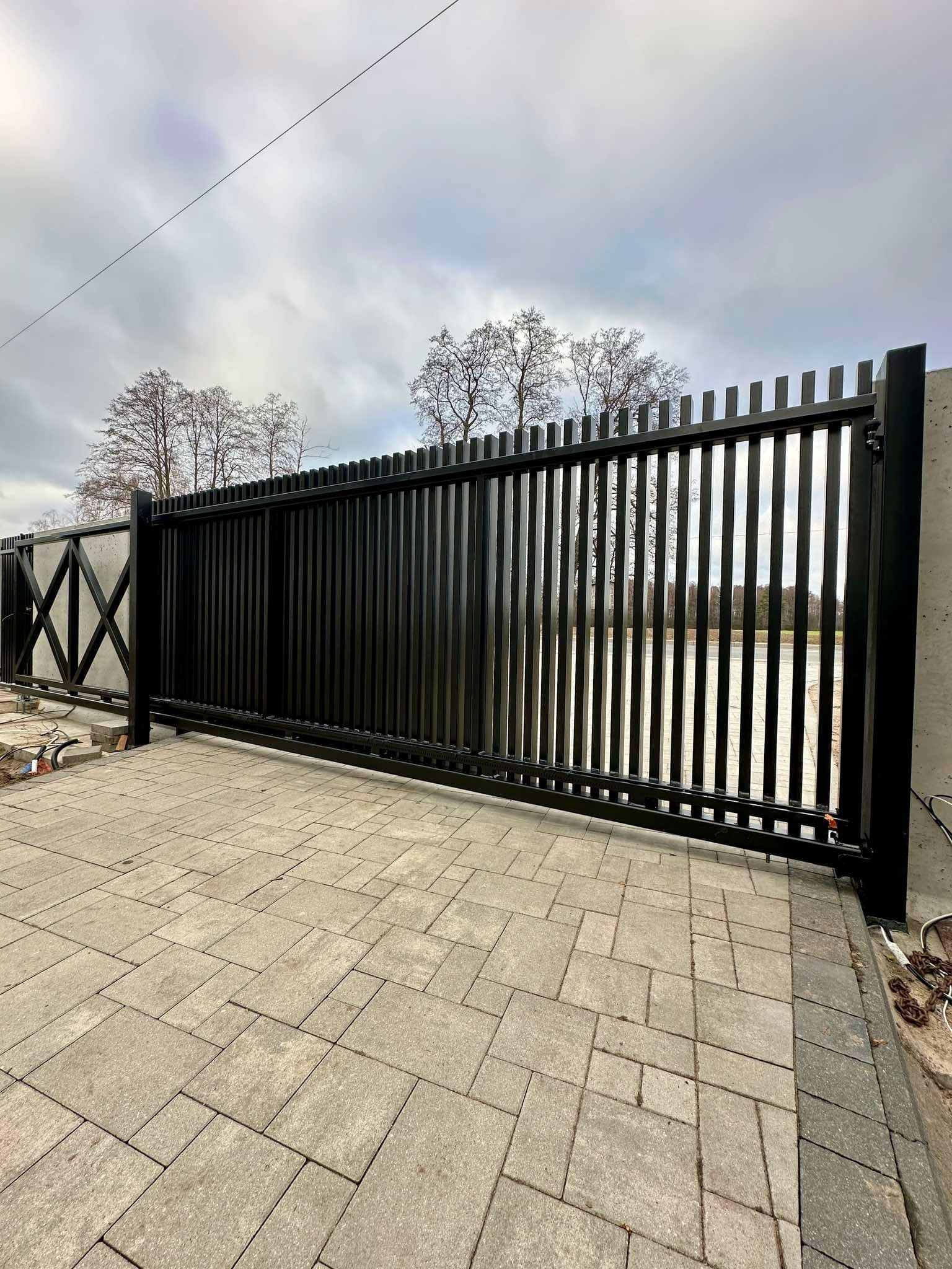 Nowoczesna, przesuwna brama wjazdowa w kolorze czarnym, z pionowymi szczeblami, zamontowana na szarej kostce brukowej, z widocznym mechanizmem jezdnym i betonowym słupkiem.